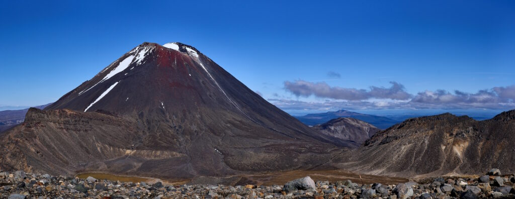 Photo du volcan néo-zélandais Ngauruhoe montrant un cône minéral noir et rouge sur lequel subsistent quelques névés.